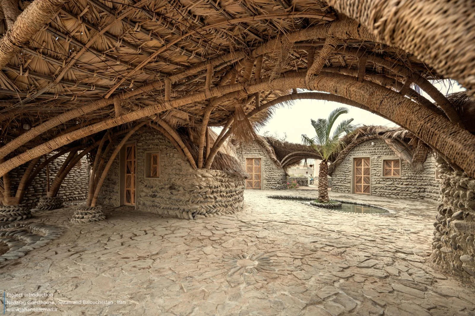 نمای خارجی و داخلی مهمانسرای ندارگ در روستای کهنانیکش، سیستان و بلوچستان، که ساختار نیمه‌باز، سقف چتری‌شکل با خرپاهای دست‌ساز و دیوارهای ضخیم گلی آن بر اساس بازتفسیر معماری بومی «کپر» و با هدف خنک‌سازی طبیعی و مشارکت جمعی اهالی طراحی و اجرا شده است.