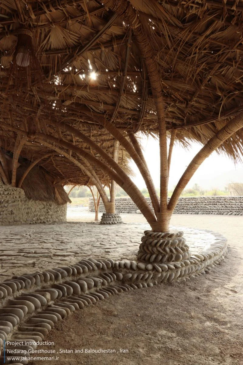 نمای خارجی و داخلی مهمانسرای ندارگ در روستای کهنانیکش، سیستان و بلوچستان، که ساختار نیمه‌باز، سقف چتری‌شکل با خرپاهای دست‌ساز و دیوارهای ضخیم گلی آن بر اساس بازتفسیر معماری بومی «کپر» و با هدف خنک‌سازی طبیعی و مشارکت جمعی اهالی طراحی و اجرا شده است.