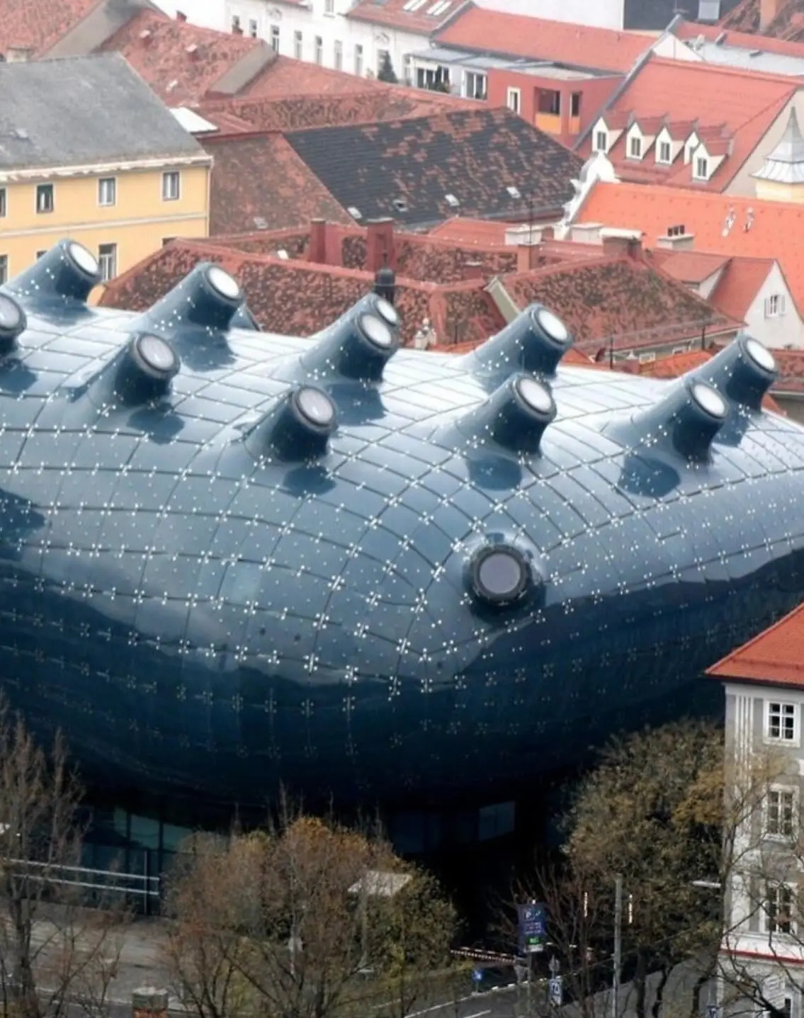 تصویری از نمای خارجی ساختمان موزه هنر کونست‌هاوس (Kunsthaus) در گراتس اتریش، که ساختار ارگانیک و غیرمعمول آن شبیه به یک موجود زنده با پوسته آبی رنگ است و به عنوان یک نماد معماری مدرن و «بیگانه دوست» طراحی شده توسط کولین فورنیر و پیتر کوک شناخته می‌شود.